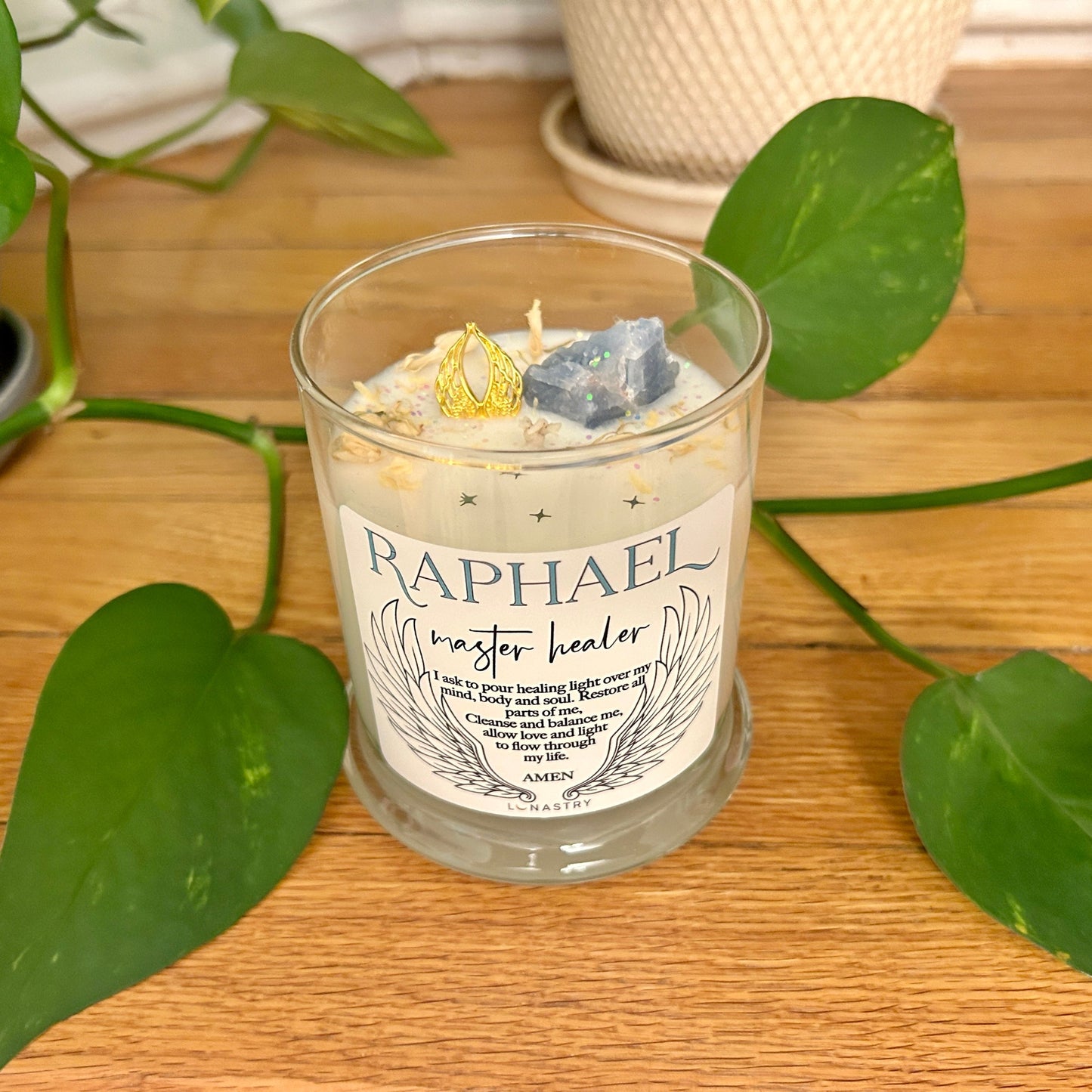 Candle in a glass jar with 'Raphael' label on a wooden surface with green leaves.