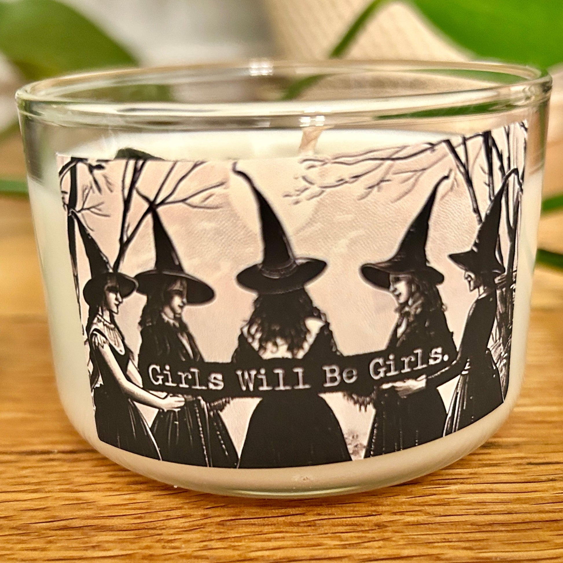 Candle in a glass holder with black and white design of women in witch hats and 'Girls Will Be Girls' text.