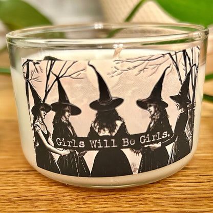 Candle in a glass holder with black and white design of women in witch hats and 'Girls Will Be Girls' text.