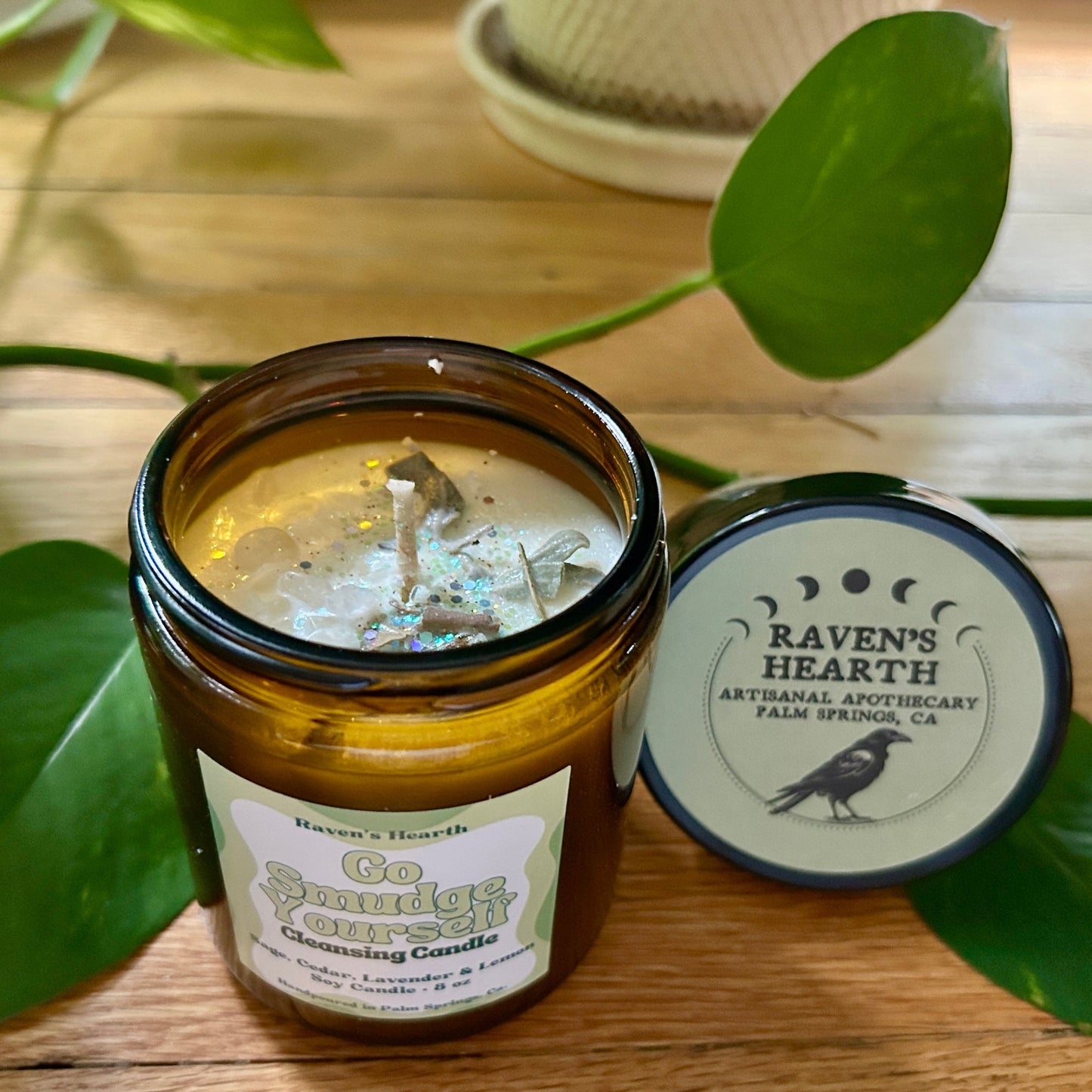 Sage cedar and lemon candle in an amber jar with a label, surrounded by green leaves on a wooden surface.