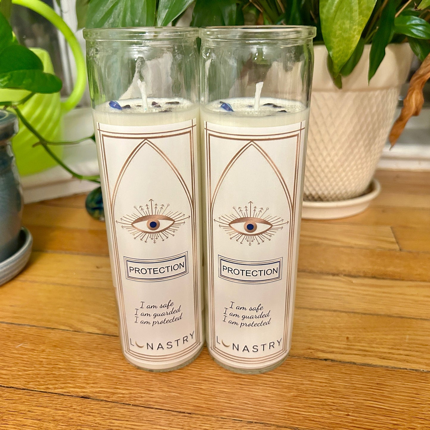 Two candles labeled 'Protection' by Lunastry on a wooden surface with plants in the background.