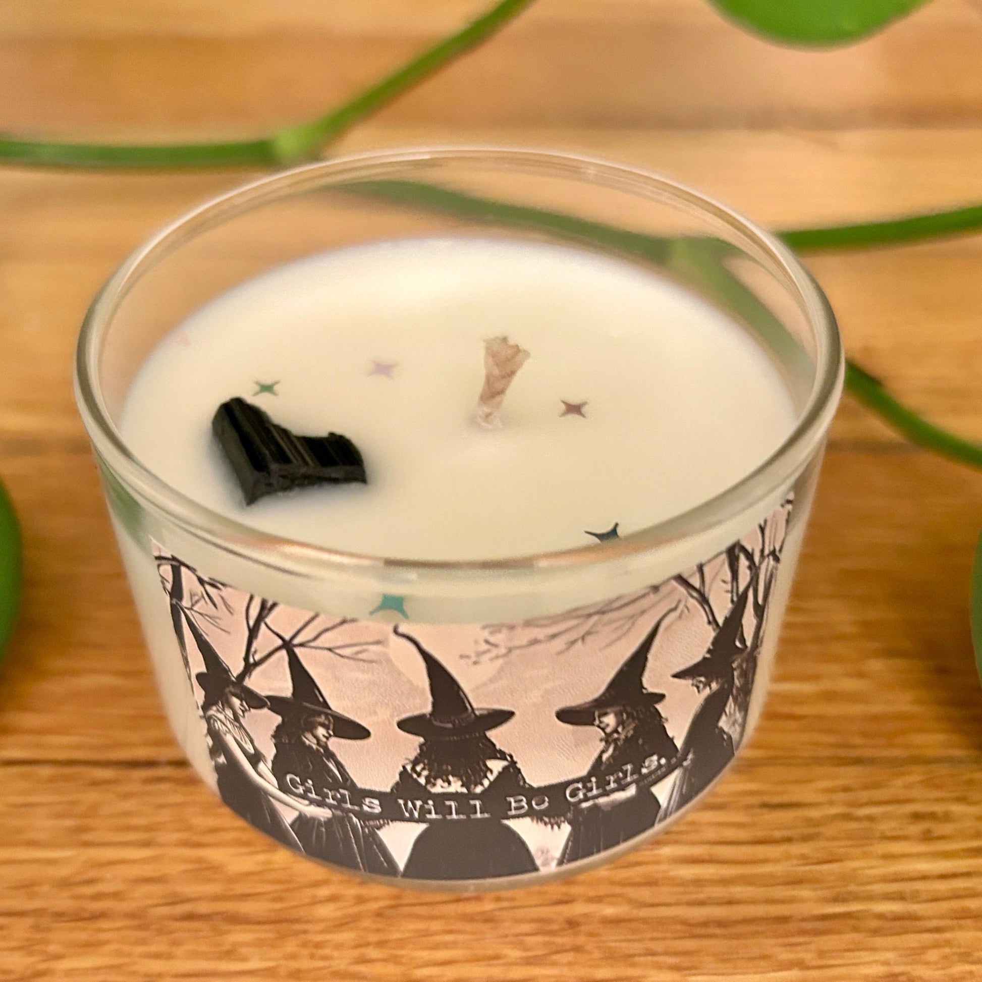 Candle in a glass jar with witch design and text on a wooden surface